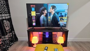 Un téléviseur posé sur un meuble, avec l’interface Netflix affichée à l’écran, deux télécommandes sur un pouf jaune et un tissu noir à motifs sur le côté gauche.