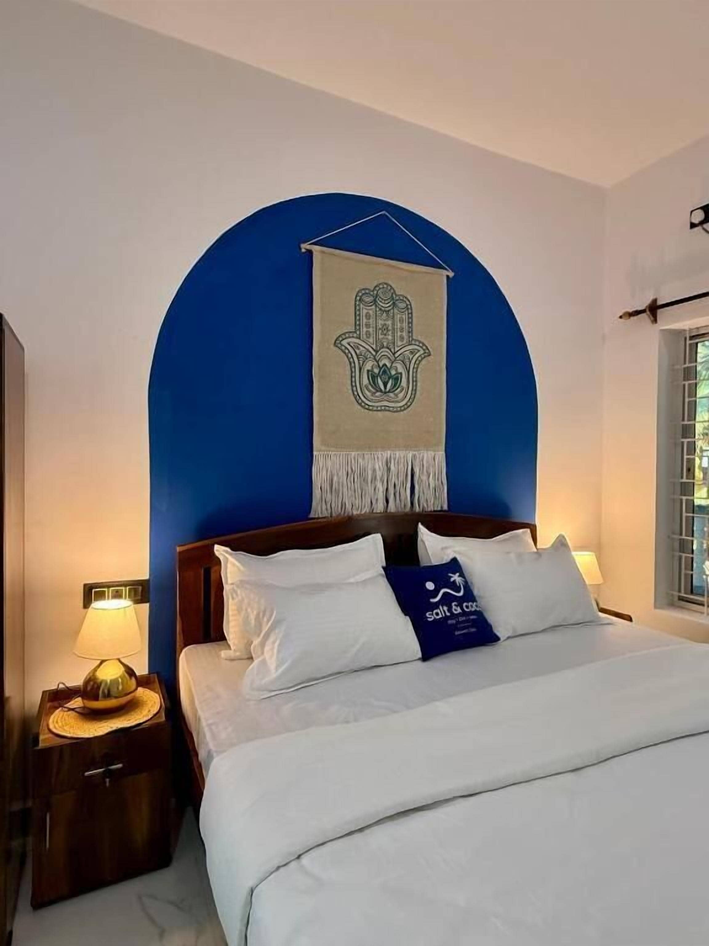 Ein Hotelzimmer mit Bett, Nachttisch, Lampe, Wandteppich und einem Fenster mit Jalousien.