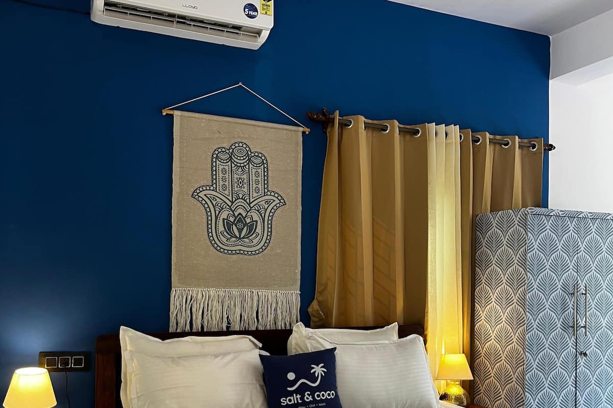 Habitación de hotel con una cama, aire acondicionado mural, un tapiz decorativo, una cortina, un papel tapiz estampado, una mesita de noche con una lámpara y una toalla enrollada sobre la cama.