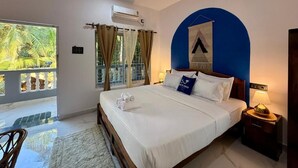A hotel room with a bed, a nightstand, a wall-mounted air conditioner, a window with curtains, and a view of palm trees. - Salt & Coco (Ashvem)