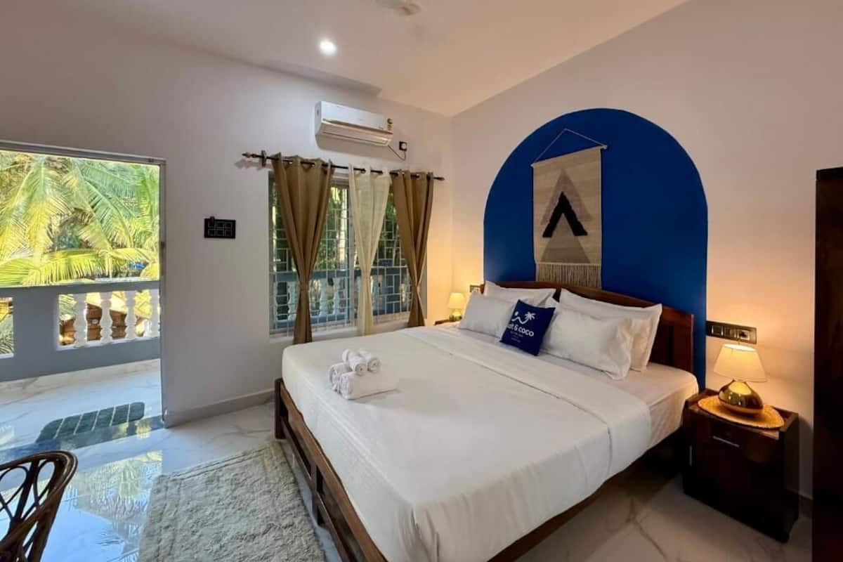 Una habitación de hotel con cama, mesita de noche, aire acondicionado de pared, ventana con cortinas y vista a palmeras.