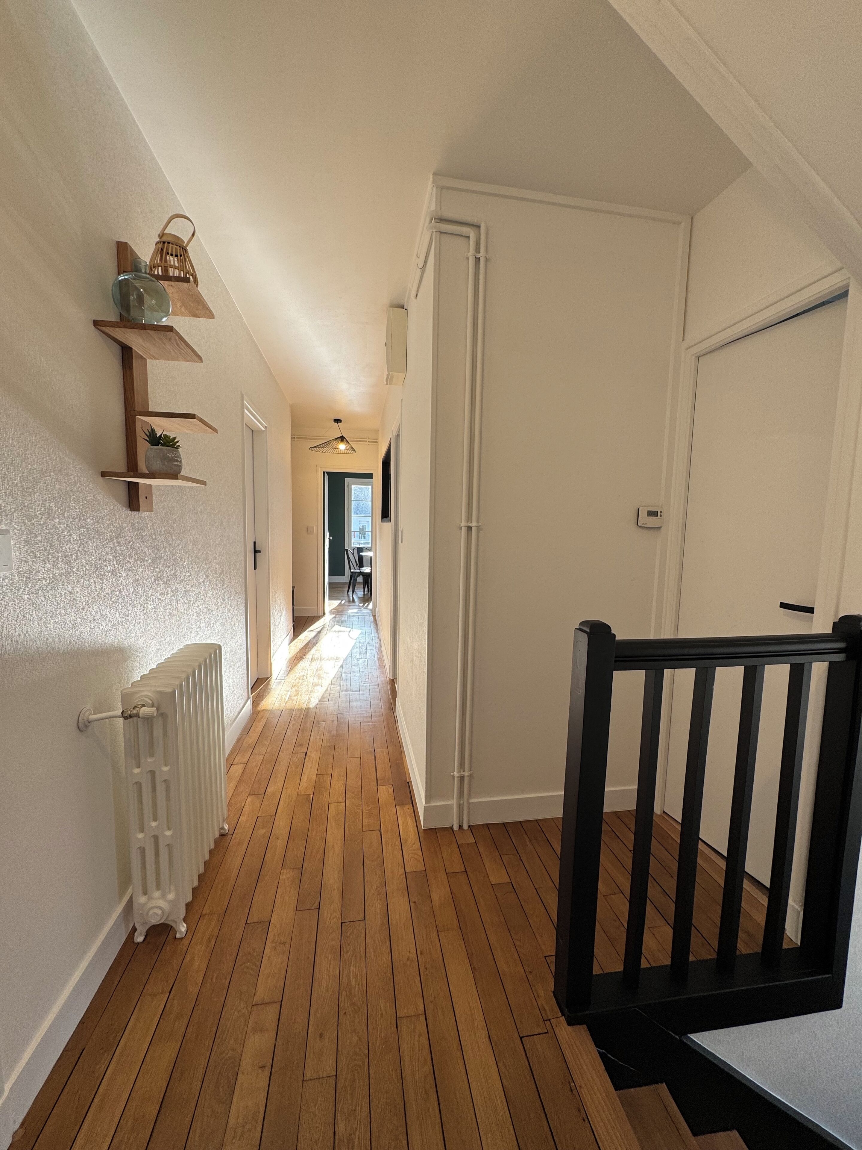 Un couloir avec un plancher en bois, des murs blancs, un radiateur, une rambarde noire et une porte au fond.