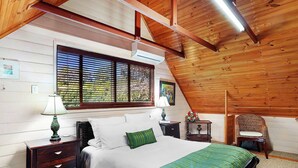 Un dormitorio con una cama, dos mesitas de noche, un aparador, una ventana con persianas, una silla y techo de madera.