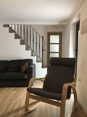Ein Wohnzimmer mit einem Holzschaukelstuhl, einem dunklen Sofa, einer Treppe mit schwarzen Geländern und einer Tür mit satinierten Glasscheiben.