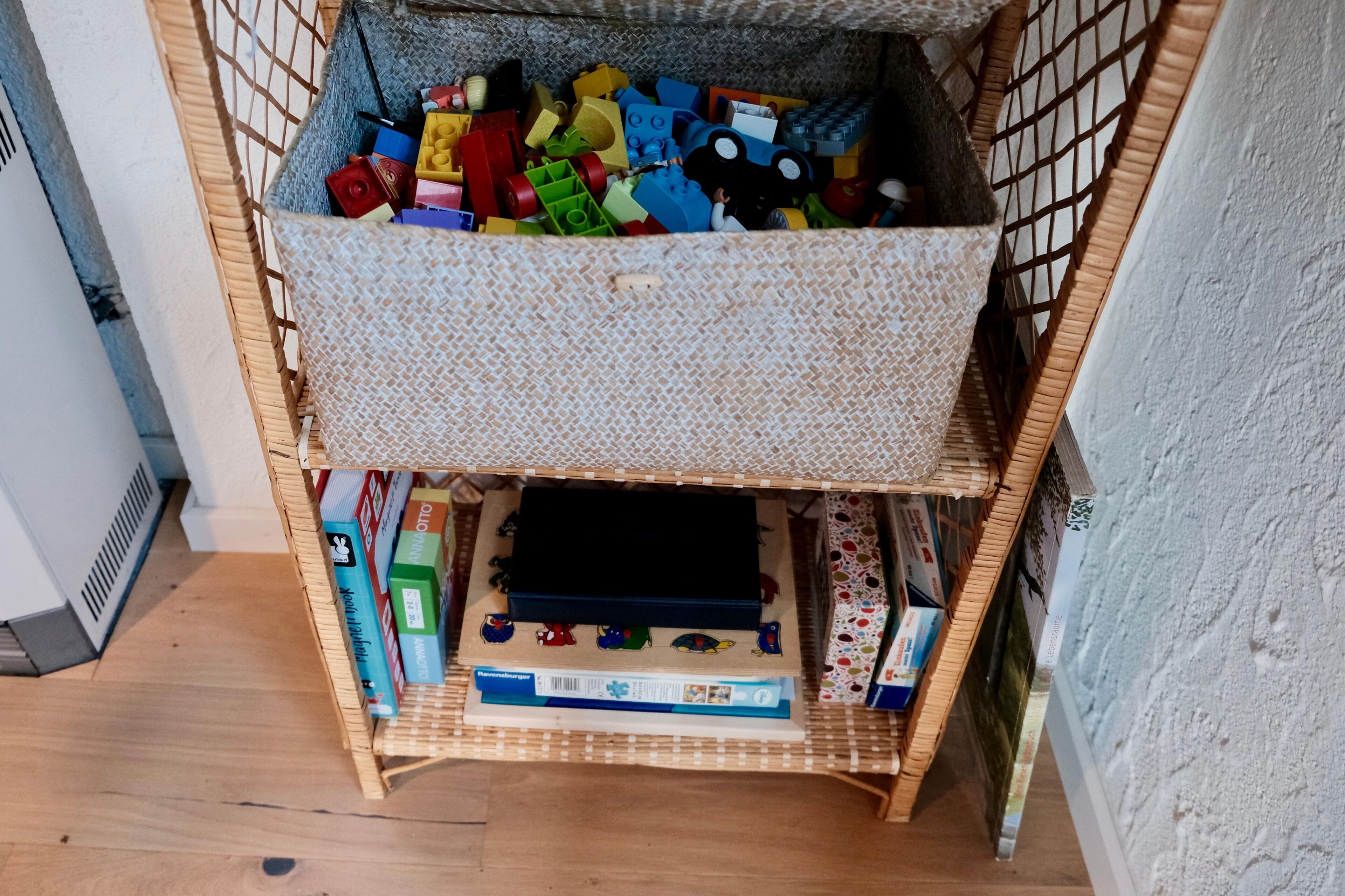 Una habitación con una estantería, una pequeña mesa de madera y un baúl lleno de objetos de colores.