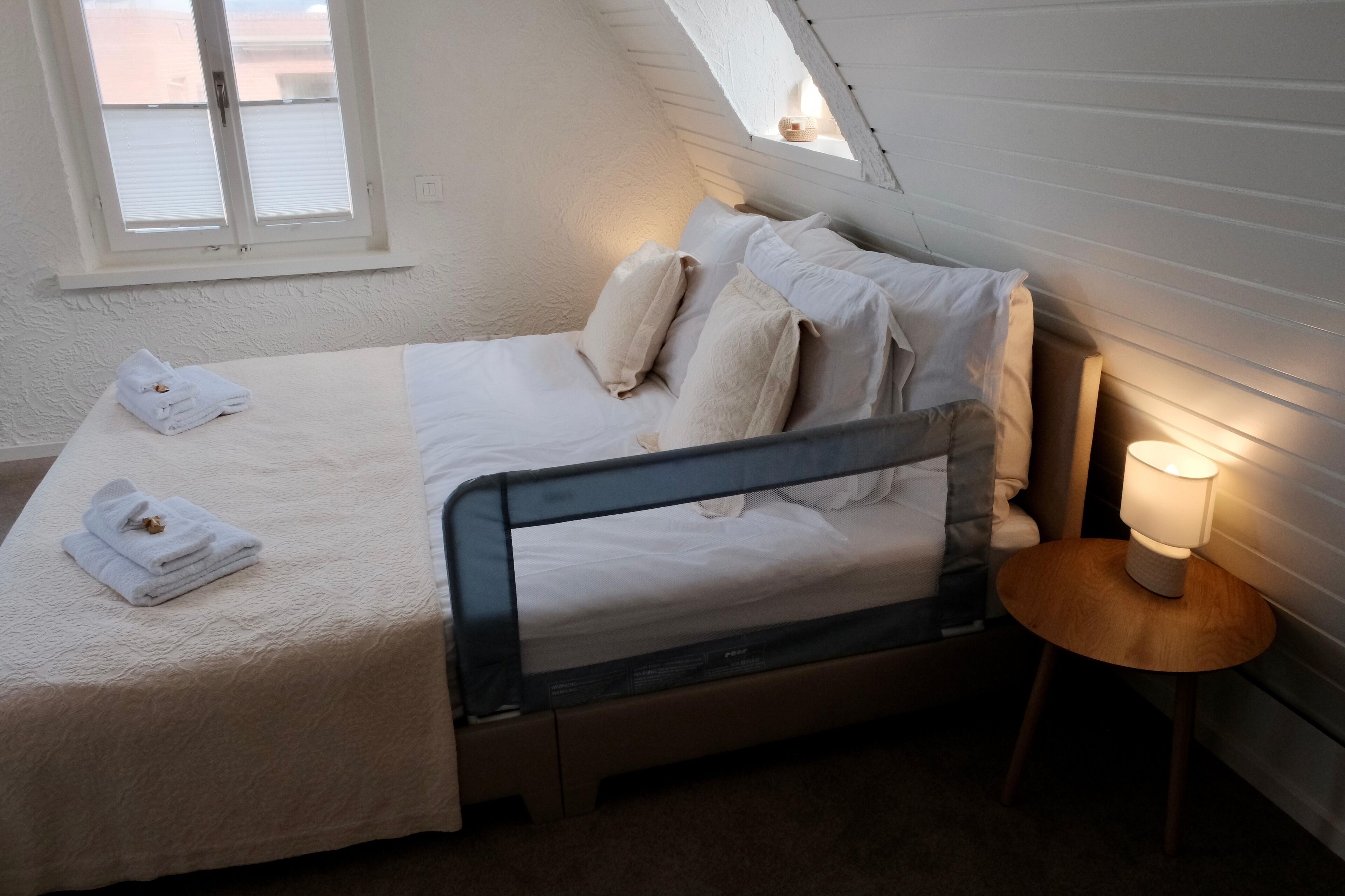 Ein ordentlich bezogenes Bett mit Kissen und einem gefalteten Handtuch, ein Nachttisch mit Lampe sowie ein Fenster mit Jalousien.