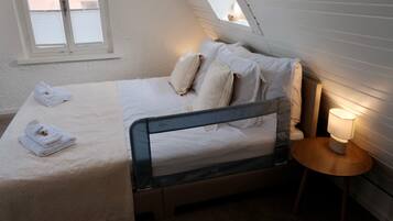 Ein ordentlich bezogenes Bett mit Kissen und einem gefalteten Handtuch, ein Nachttisch mit Lampe sowie ein Fenster mit Jalousien.