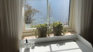 Een raam met uitzicht op een meer en bomen, een wit bureau met een plant, en een beker met rietje.