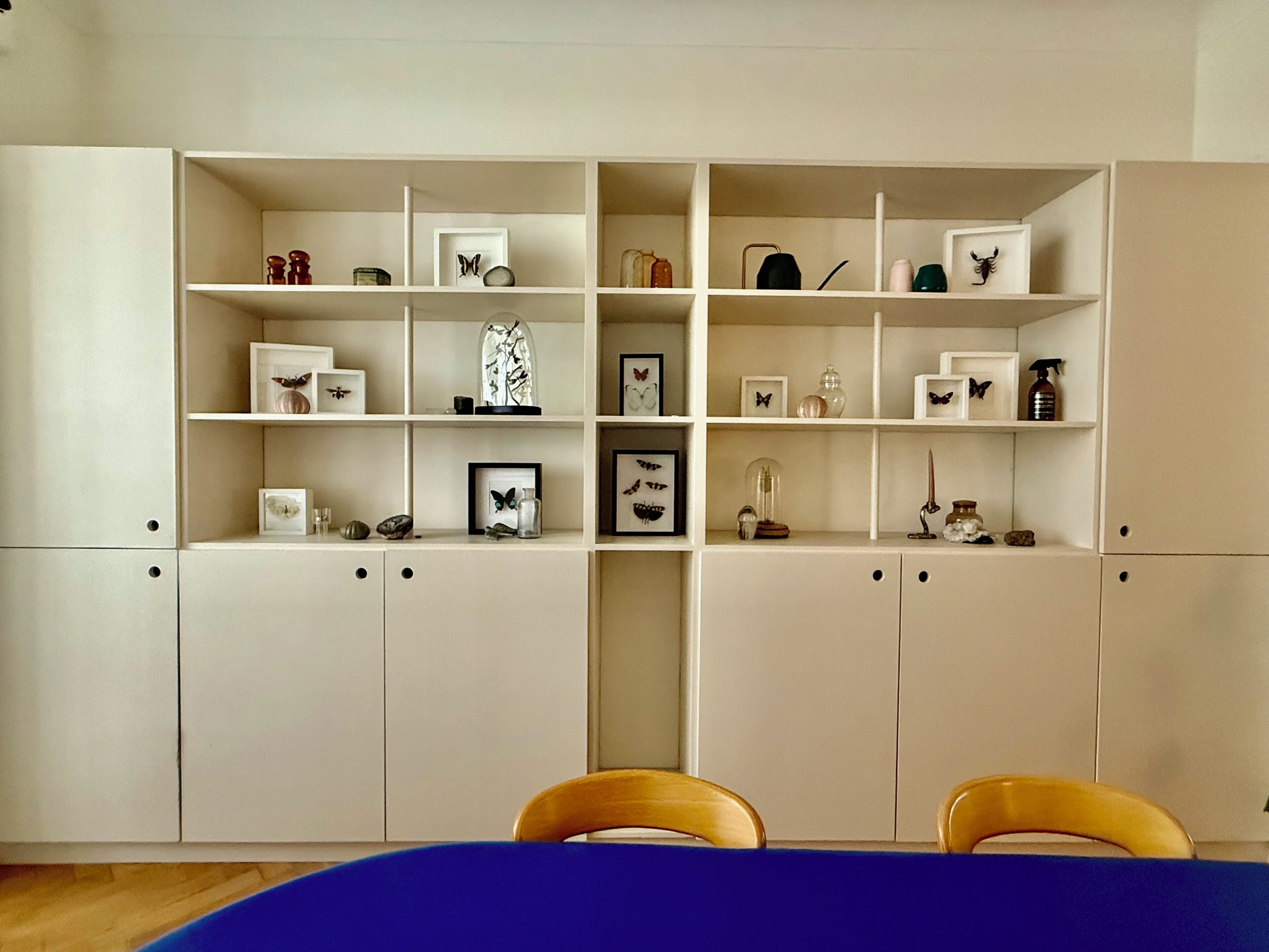 A room with a white cabinet with multiple shelves, a blue table, and two wooden chairs.