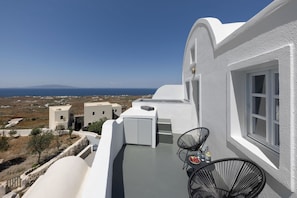 A white building with a balcony, a table, two chairs, and a view of the sea and distant buildings. - Elios Cave Suites (Oia)