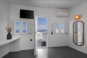 A modern room with a large TV, two windows with shutters, an open door leading to a balcony, a mirror, and an air conditioning unit. - Elios Cave Suites (Oia)