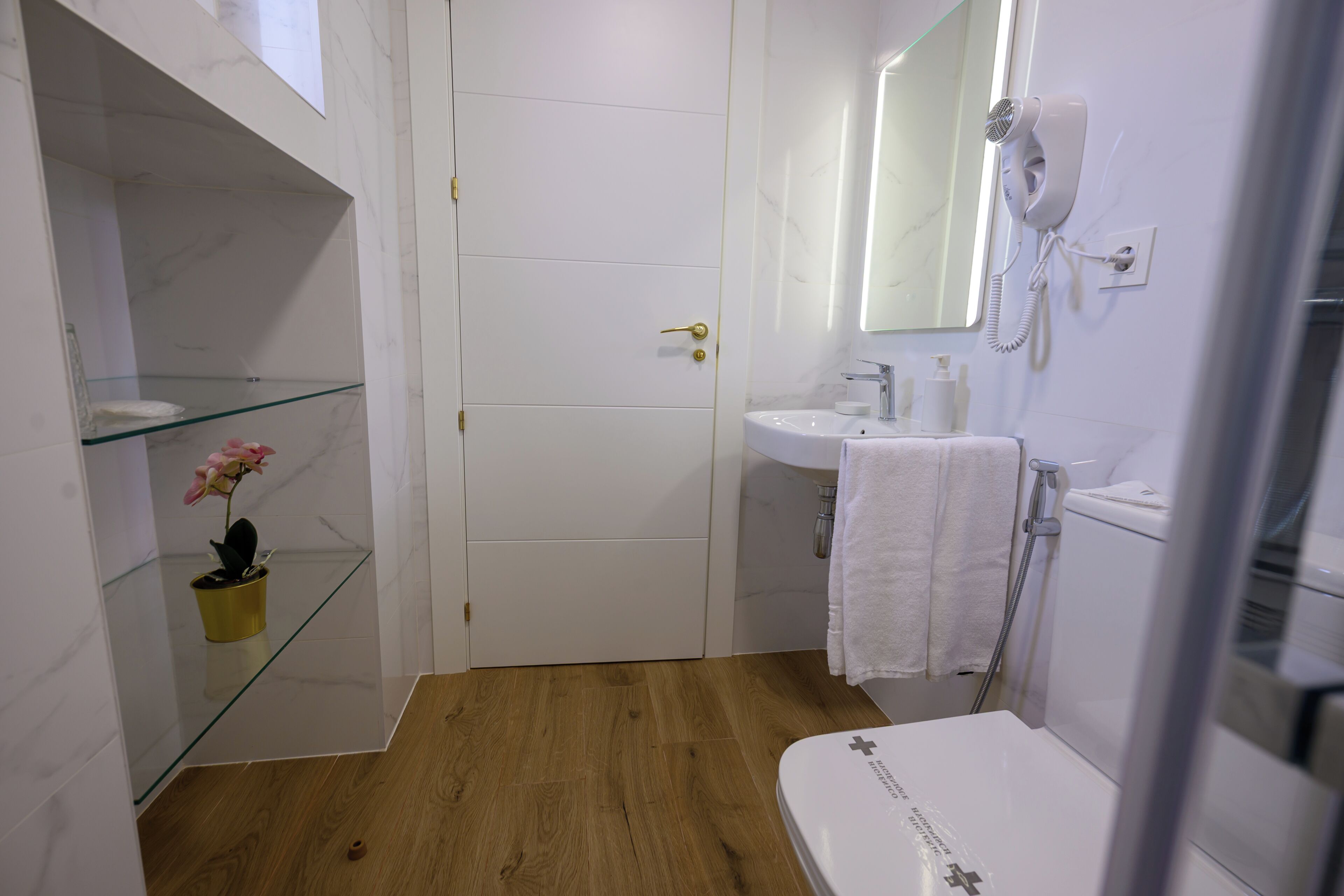 A bathroom with a white toilet, a glass shelf with a potted plant, a sink with a towel, and a hairdryer mounted on the wall.