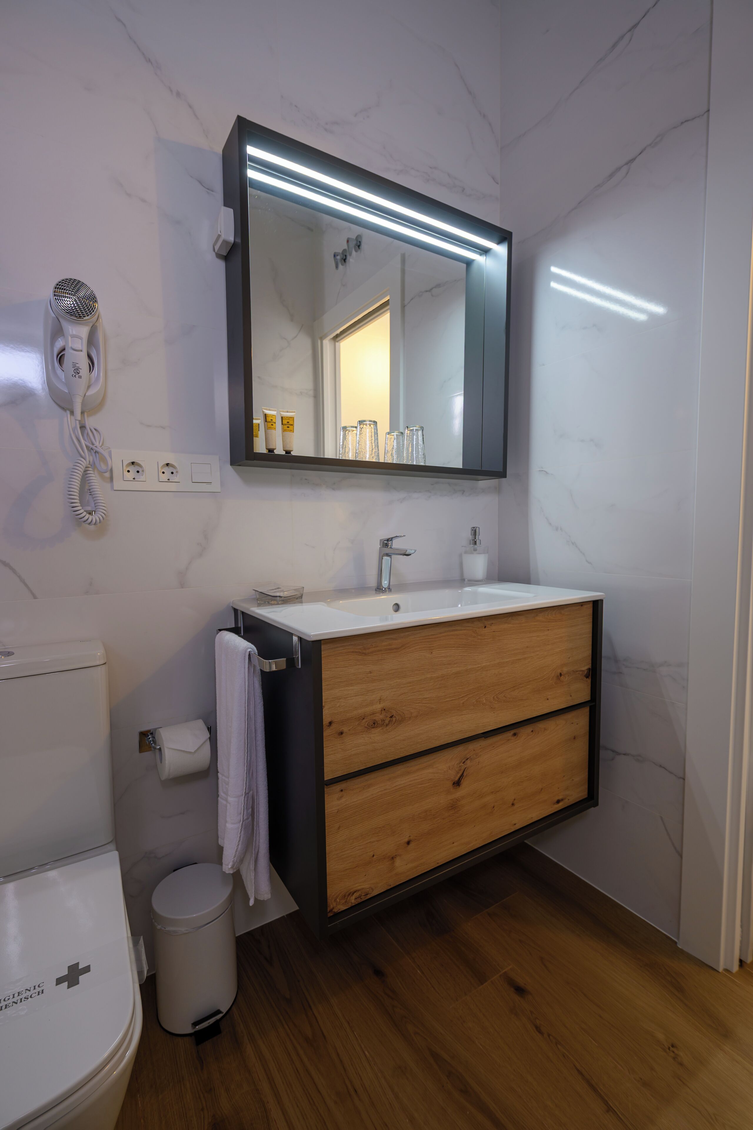 Un baño moderno con pared de mármol, tocador de madera, un espejo grande con iluminación debajo del mueble, un secador de pelo y un inodoro con un signo '+'.