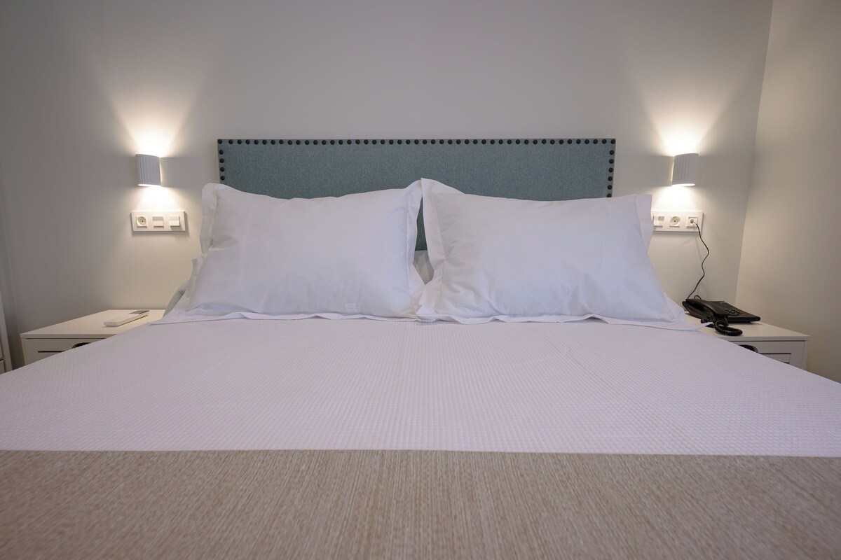 A neatly made bed with white linens, two bedside lamps, a telephone, and a wall socket.