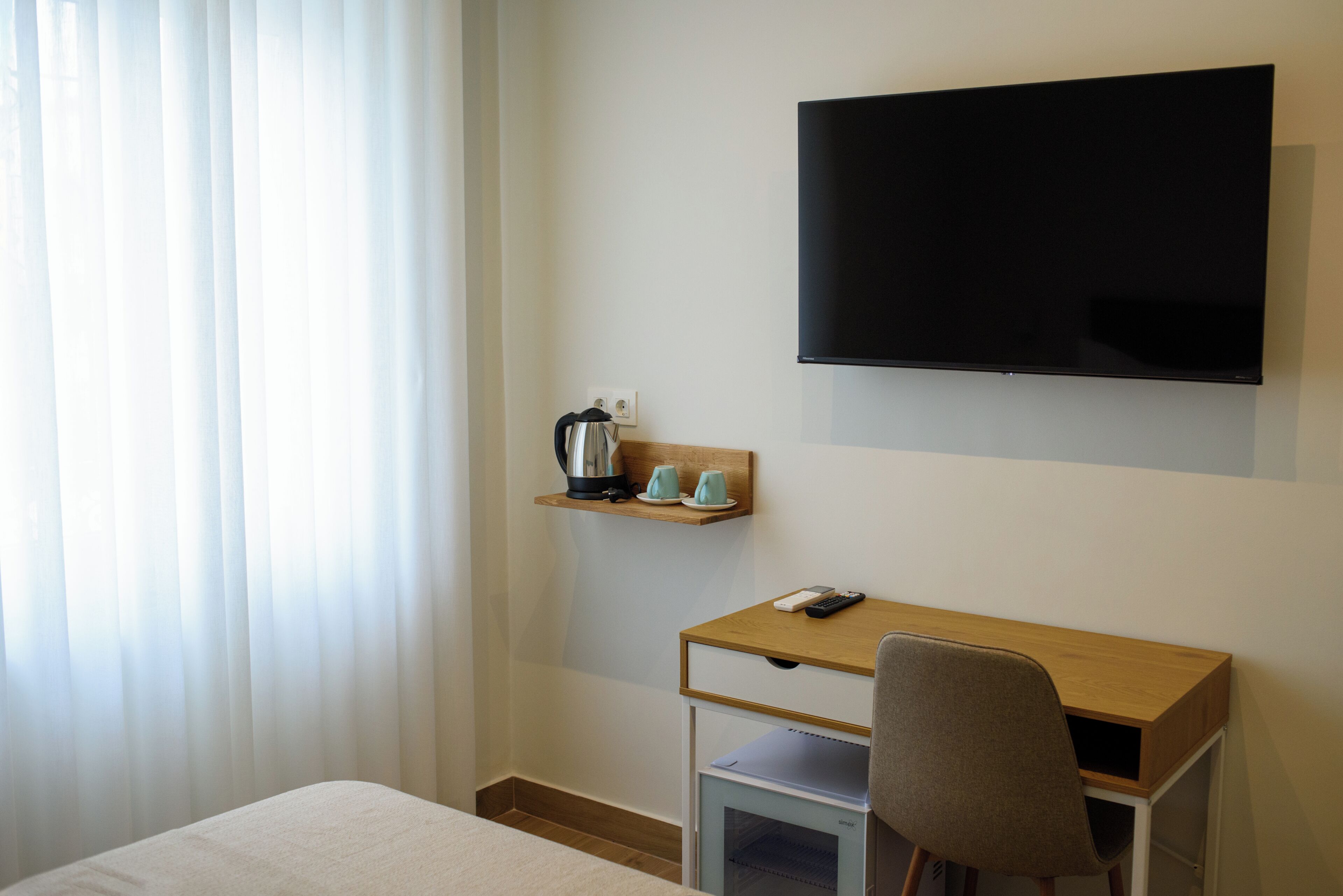 Ein Hotelzimmer mit einem Bett, einem Schreibtisch mit Stuhl, einem Fernseher und einem Regal mit Wasserkocher und Tassen.