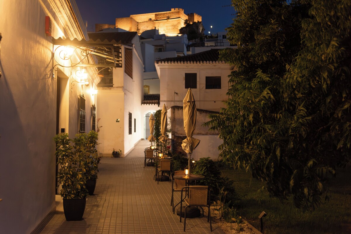 A narrow alleyway with a row of outdoor seating, potted plants, and a building with a balcony.