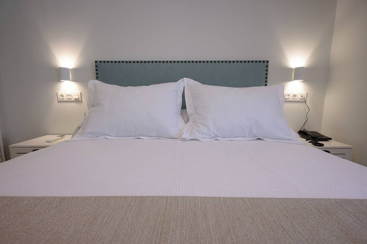 A neatly made bed with white linens, two bedside lamps, a telephone, and a wall socket.