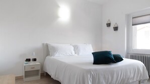 Un lit blanc avec deux oreillers, une table de chevet avec une lampe et une fenêtre avec des stores.
