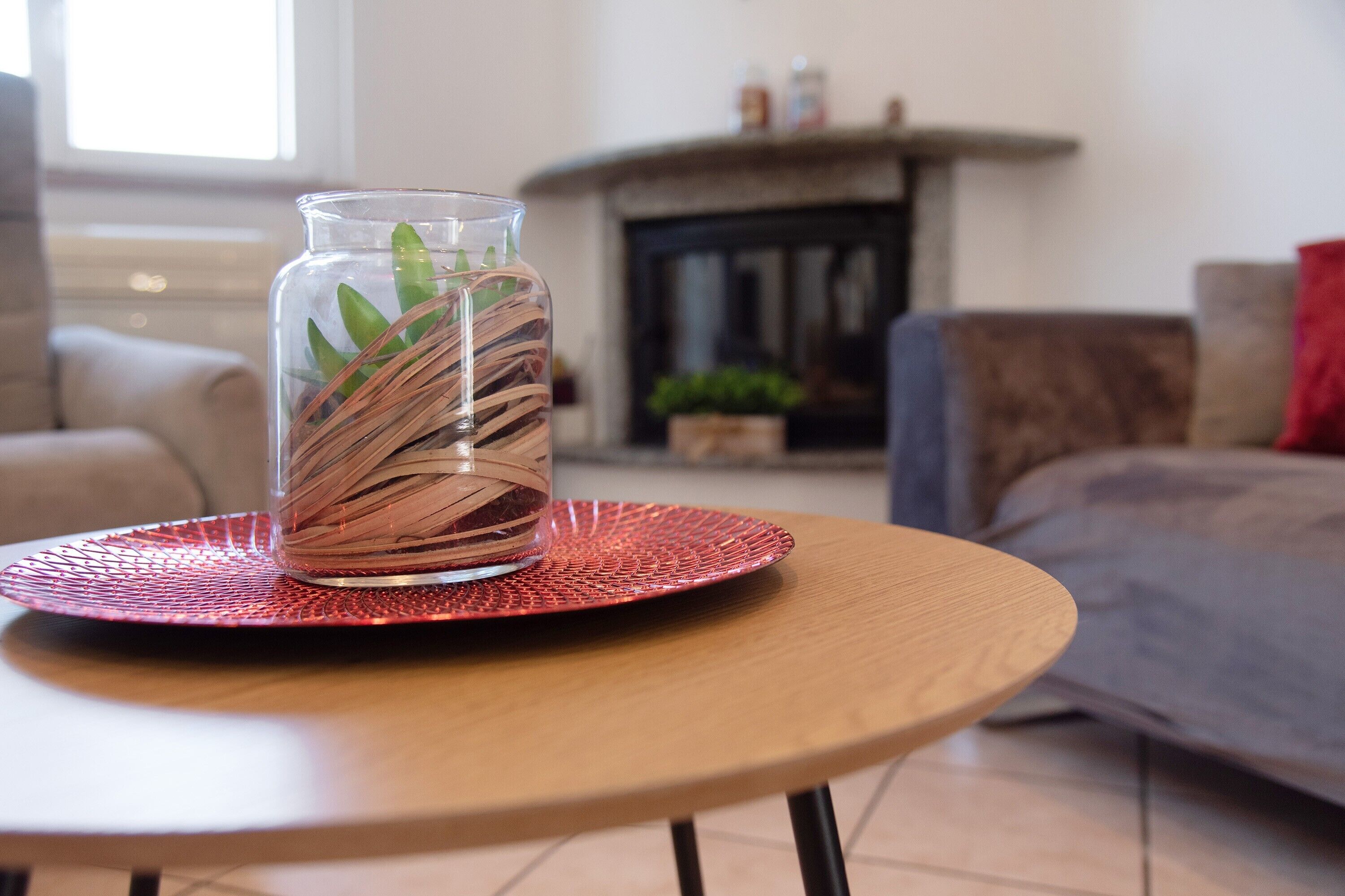 A glass vase with green leaves on a wooden table with a red placemat, in a room with a fireplace and a sofa.