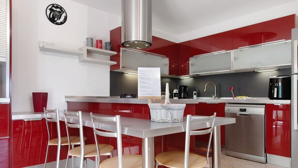 Une cuisine moderne avec des meubles de couleur rouge, des appareils en acier inoxydable et une table à manger blanche, accompagnée de quatre chaises.