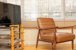 Ein Zimmer mit einem Fernseher auf einer Glasablage, einem Ledersessel und einem Fenster mit Jalousien.