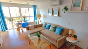Una sala de estar con un sofá, una mesa de centro de vidrio, una mesita de noche con una lámpara y una zona de comedor con una mesa y sillas.