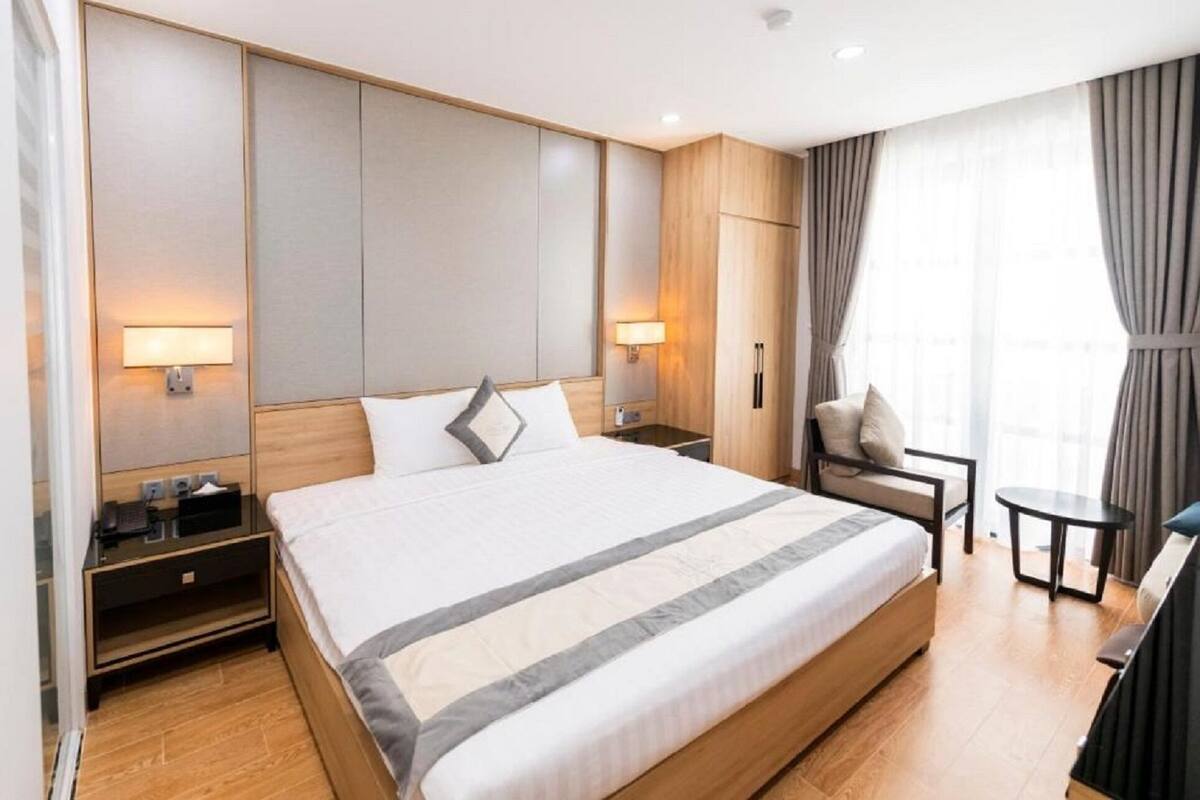 A hotel room with a large bed, bedside tables, a chair, a small table, and a window with curtains.