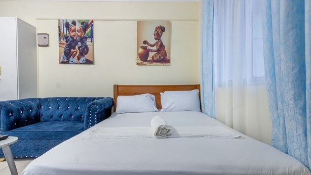 Une chambre d’hôtel avec un lit, un canapé bleu, deux tableaux accrochés au mur et une fenêtre avec des rideaux légers.