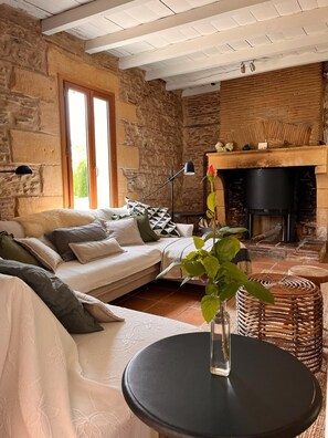 A cozy living room with a stone wall, a fireplace, a sofa, and a round table with a vase. - Chez le Vigneron | Tasteful and elegant vacation home with pool in the Dordogne (Monfaucon)