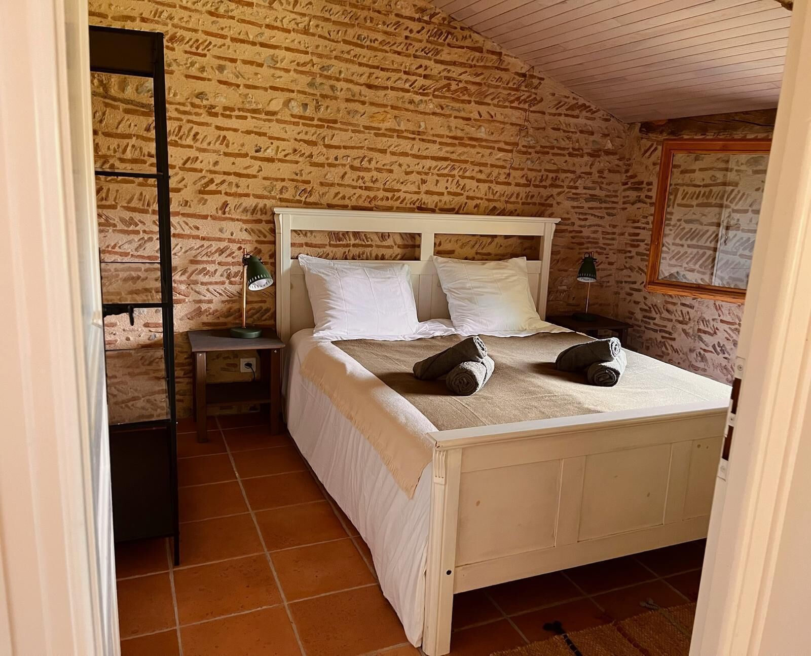 Une chambre avec un mur décoré d’un motif en briques, un lit en bois avec une literie blanche, deux lampes de chevet et une petite table de nuit.