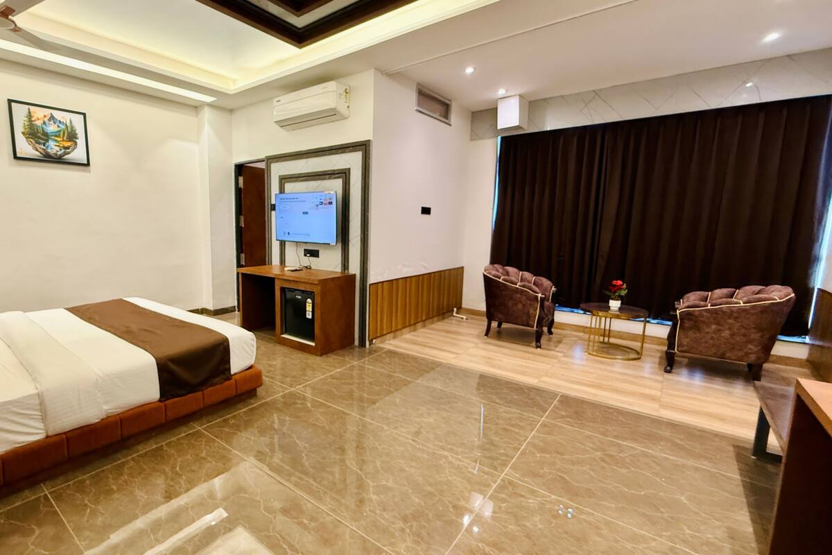 Una camera d'albergo con un letto, una scrivania con un computer, due poltrone, una TV e un'ampia finestra con tende.