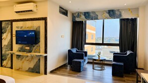 Une chambre d’hôtel moderne avec un mur d’accent en marbre, un téléviseur fixé au mur, deux fauteuils, une petite table ronde et une fenêtre donnant sur la ville.