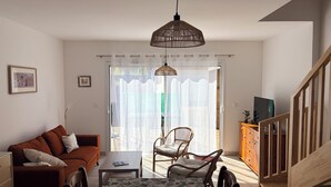 A living room with a sofa, a coffee table, and a dining table with chairs. There is a staircase to the right and a door leading outside. - DUPLEX house in a Landes village near the ocean (Lit-et-Mixe)