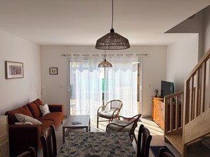 Ein Wohnzimmer mit einer Couch, einem Couchtisch und einem Esstisch mit Stühlen. Eine Treppe befindet sich rechts und eine Tür führt nach draußen.