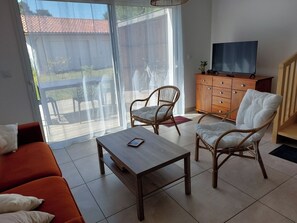A living room with a sofa, two wicker chairs, a coffee table, a wooden cabinet with a television, and a sliding glass door leading to an outdoor area. - DUPLEX house in a rural village near the ocean (Lit-et-Mixe)
