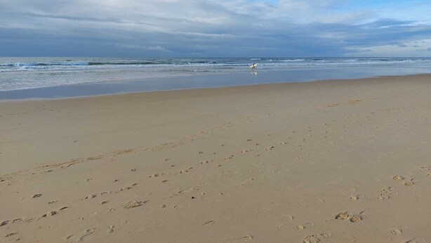 A beach with footprints leading to the water, a person walking with a surfboard, and a cloudy sky. - DUPLEX house in a Landes village near the ocean (Lit-et-Mixe)