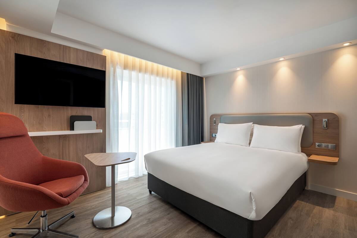 A modern hotel room with a large bed, a red armchair, a small table, a TV mounted on the wall, and a window with curtains.