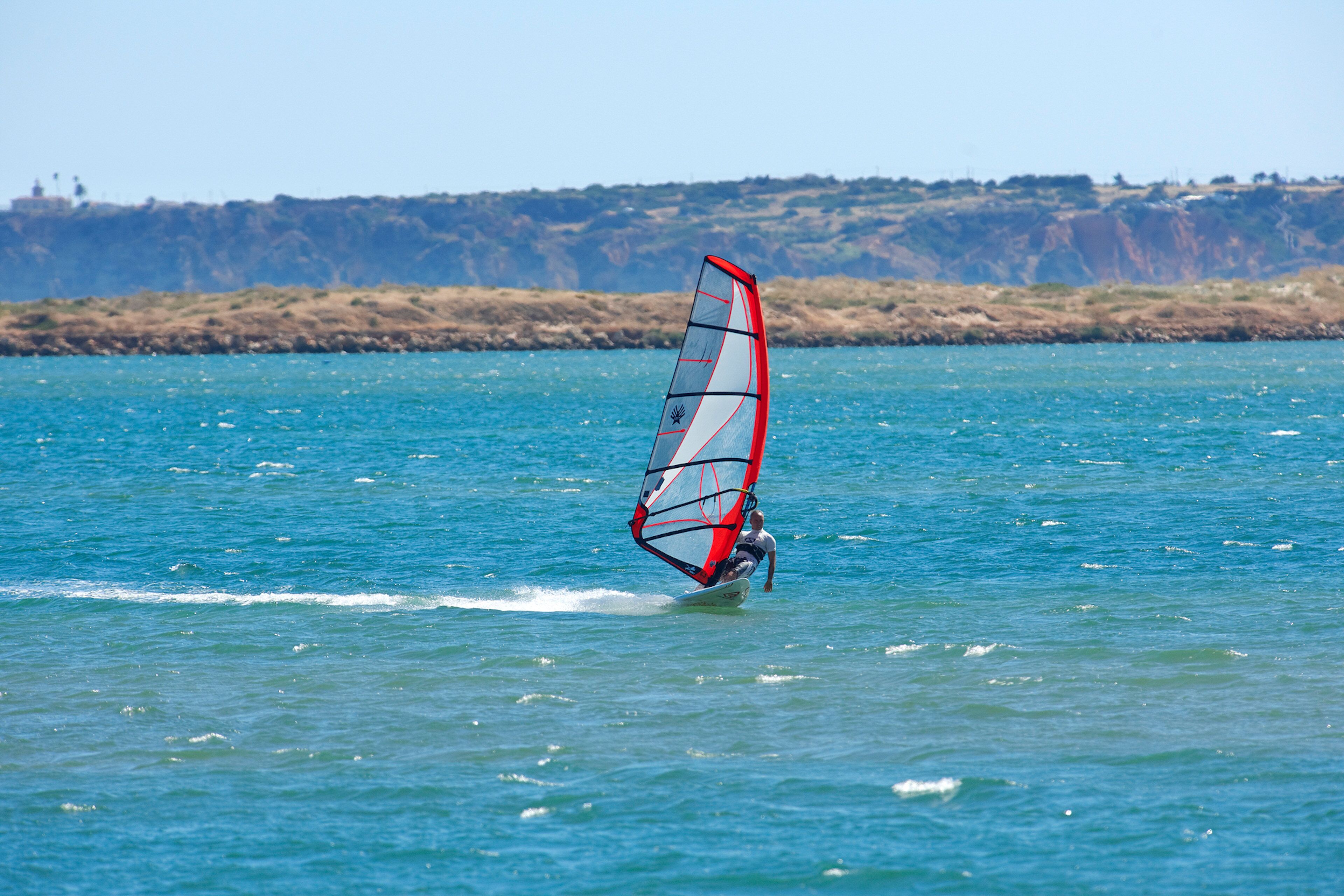 A person windsurfing on a calm sea with a clear sky and a rocky coastline in the background.