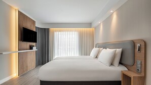 Et moderne hotelværelse med en stor seng, et fladskærms-TV monteret på en træpanel, et sengebord med en lampe og et stort vindue med lette gardiner.