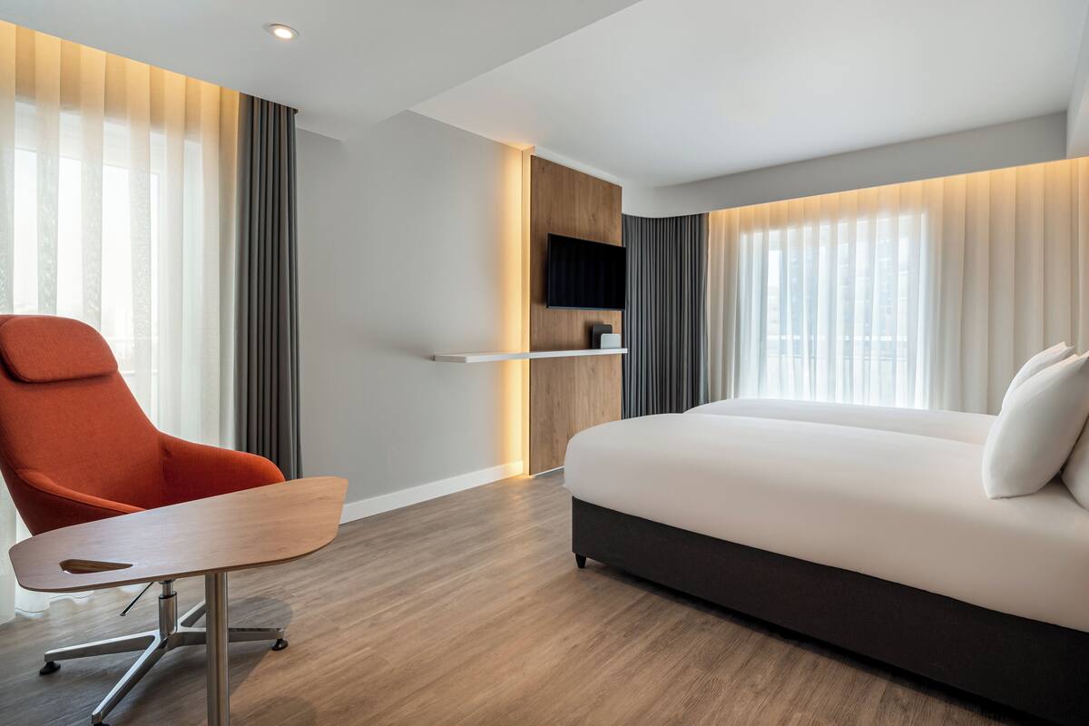 A modern hotel room with a wooden floor, a sofa, an orange chair, a TV mounted on a wall, and a window with curtains.