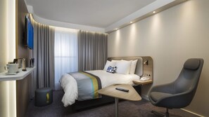 Ein Hotelzimmer mit Bett, Nachttisch, Stuhl und einem kleinen Schreibtisch mit Fernbedienung.