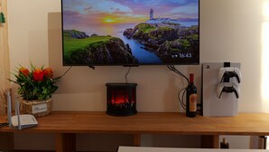 Un téléviseur à écran plat fixé au mur, diffusant une image d’un magnifique coucher de soleil, une bouteille de vin, une console de jeux et une petite table sur laquelle est posé un vase de fleurs.
