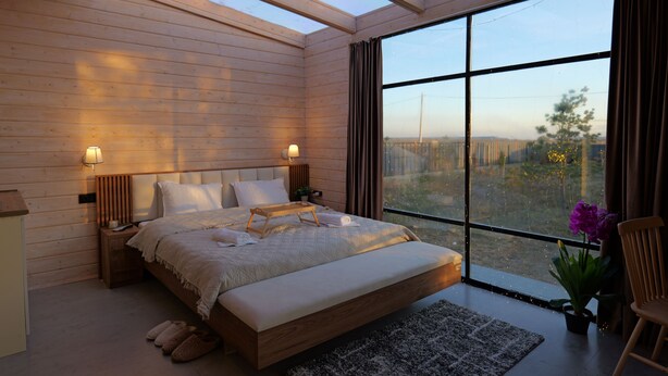 Une chambre avec une grande fenêtre, des murs en bois, un lit avec des oreillers, une table de chevet et une commode.
