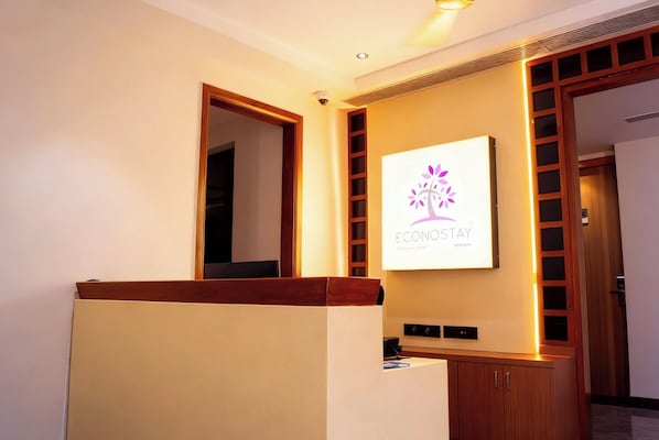 A reception area with a wooden reception desk, a wall-mounted sign with a logo, and a door with a wooden frame. - Econostay (Bhilwara)