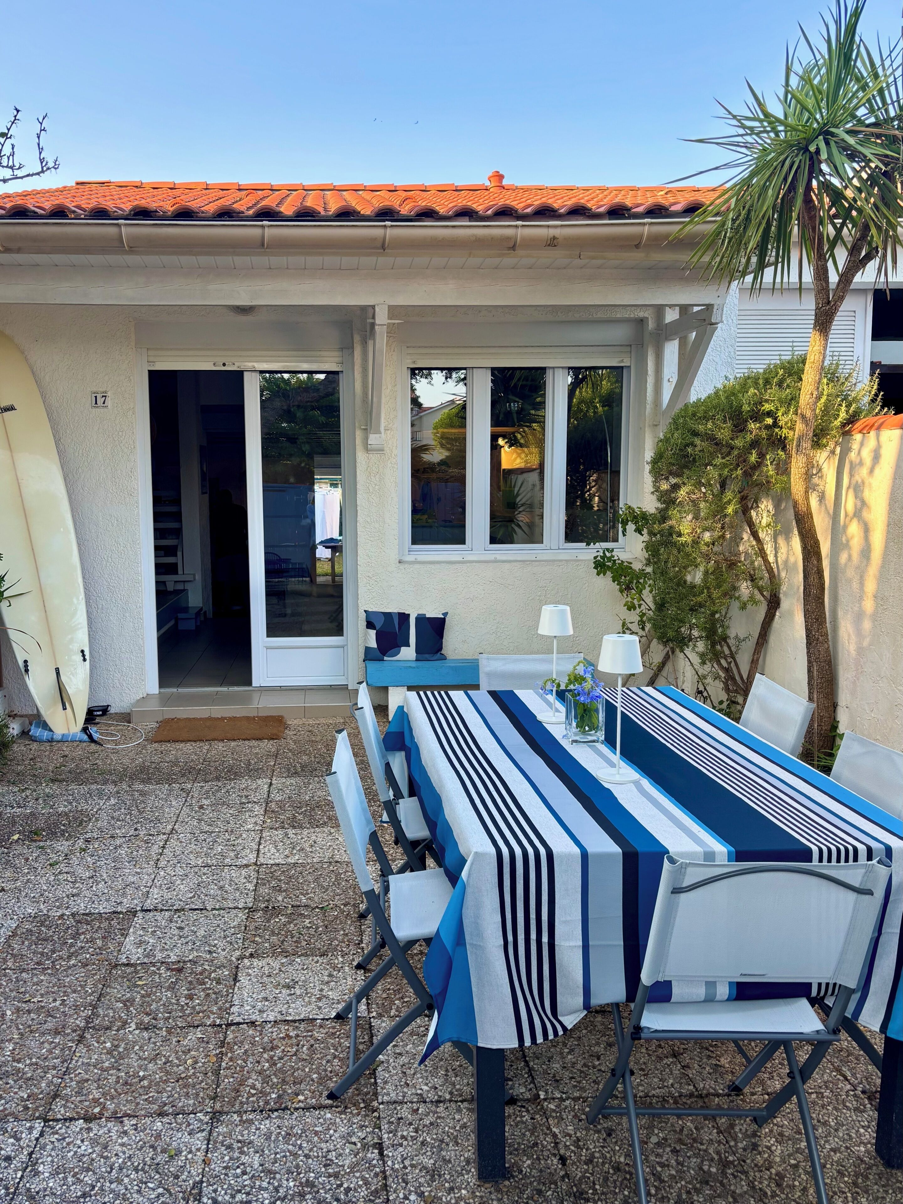 An outdoor dining area with a table set for two, a surfboard leaning against the wall, and a palm tree.