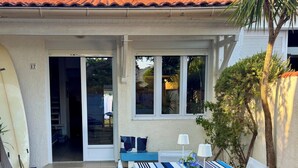 An outdoor dining area with a table set for two, a surfboard leaning against the wall, and a palm tree. - Villa Olinka, behind the dune, plage des Bourdaines (Seignosse)