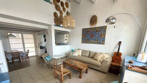 A living room with a sofa, coffee table, and chairs, a dining area with a table and chairs, a kitchen area, and a guitar. - Villa Olinka, behind the dune, plage des Bourdaines (Seignosse)