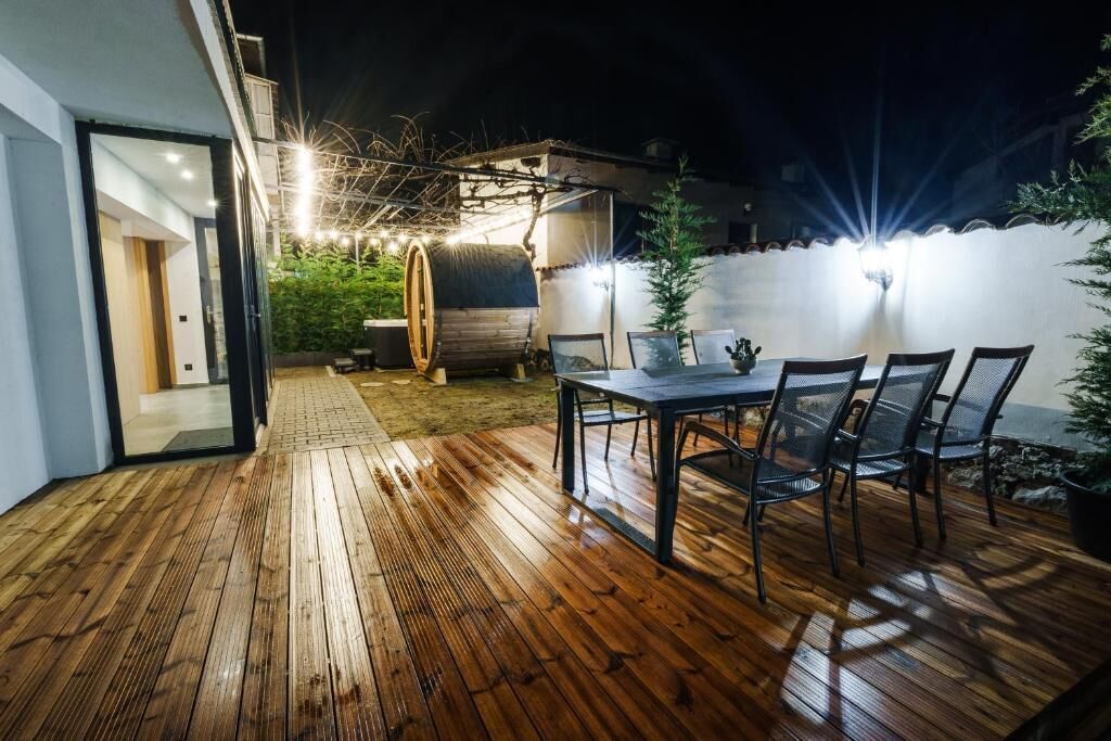 Eine Außenpatio mit Holzdeck, einem Esstisch mit Stühlen, einem Fass und einem kleinen Baum.