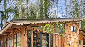 A wooden cabin with a gravel driveway, a red chair outside, and a small tree in front. - Unique Log cabin @ Rainier with Hot Tub-Fast WiFi (Ashford)