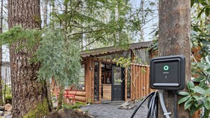 A rustic cabin with a gravel driveway, a wooden door, and a charging station attached to a tree. - Unique Log cabin @ Rainier with Hot Tub-Fast WiFi (Ashford)